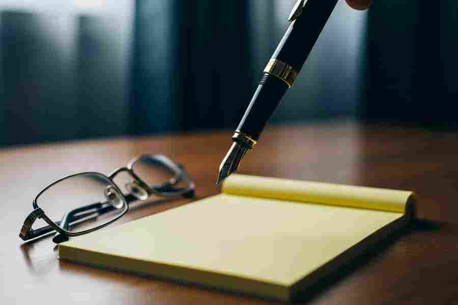 Fountain pen poised above a blank notepad with reading glasses on a wooden surface